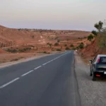 A narrow asphalt road winding through the sandy desert landscape of Morocco on a hot sunny summer day, a clear blue sky.