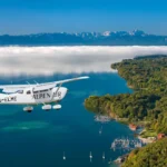 Ein ALPEN AIR Flugzeug im Flug über einem bayerischen See mit bewaldetem Ufer, Yachthafen und Alpenpanorama im Hintergrund. Das Motiv verbindet Luftfahrt, Seenlandschaft und die Nähe zwischen München, Bayern und den Alpen. Es eignet sich besonders für Themen rund um Scenic Flights, Rundflüge in Bayern, Alpenrundflüge, Reiseerlebnisse, Luftaufnahmen und besondere Ausflüge im süddeutschen Alpenvorland.