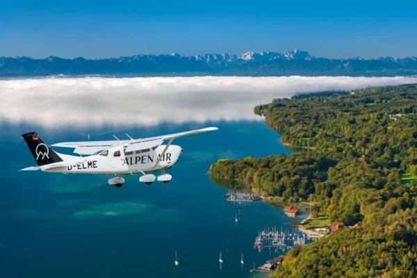 Ein ALPEN AIR Flugzeug im Flug über einem bayerischen See mit bewaldetem Ufer, Yachthafen und Alpenpanorama im Hintergrund. Das Motiv verbindet Luftfahrt, Seenlandschaft und die Nähe zwischen München, Bayern und den Alpen. Es eignet sich besonders für Themen rund um Scenic Flights, Rundflüge in Bayern, Alpenrundflüge, Reiseerlebnisse, Luftaufnahmen und besondere Ausflüge im süddeutschen Alpenvorland.