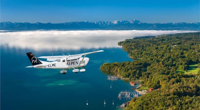 Ein ALPEN AIR Flugzeug im Flug über einem bayerischen See mit bewaldetem Ufer, Yachthafen und Alpenpanorama im Hintergrund. Das Motiv verbindet Luftfahrt, Seenlandschaft und die Nähe zwischen München, Bayern und den Alpen. Es eignet sich besonders für Themen rund um Scenic Flights, Rundflüge in Bayern, Alpenrundflüge, Reiseerlebnisse, Luftaufnahmen und besondere Ausflüge im süddeutschen Alpenvorland.
