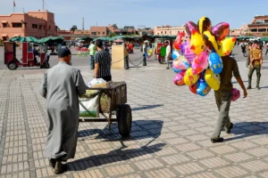Daily life scene at Jemaa el-Fnaa square in Marrakech with a local man pushing a cart and a street vendor carrying colorful balloons.